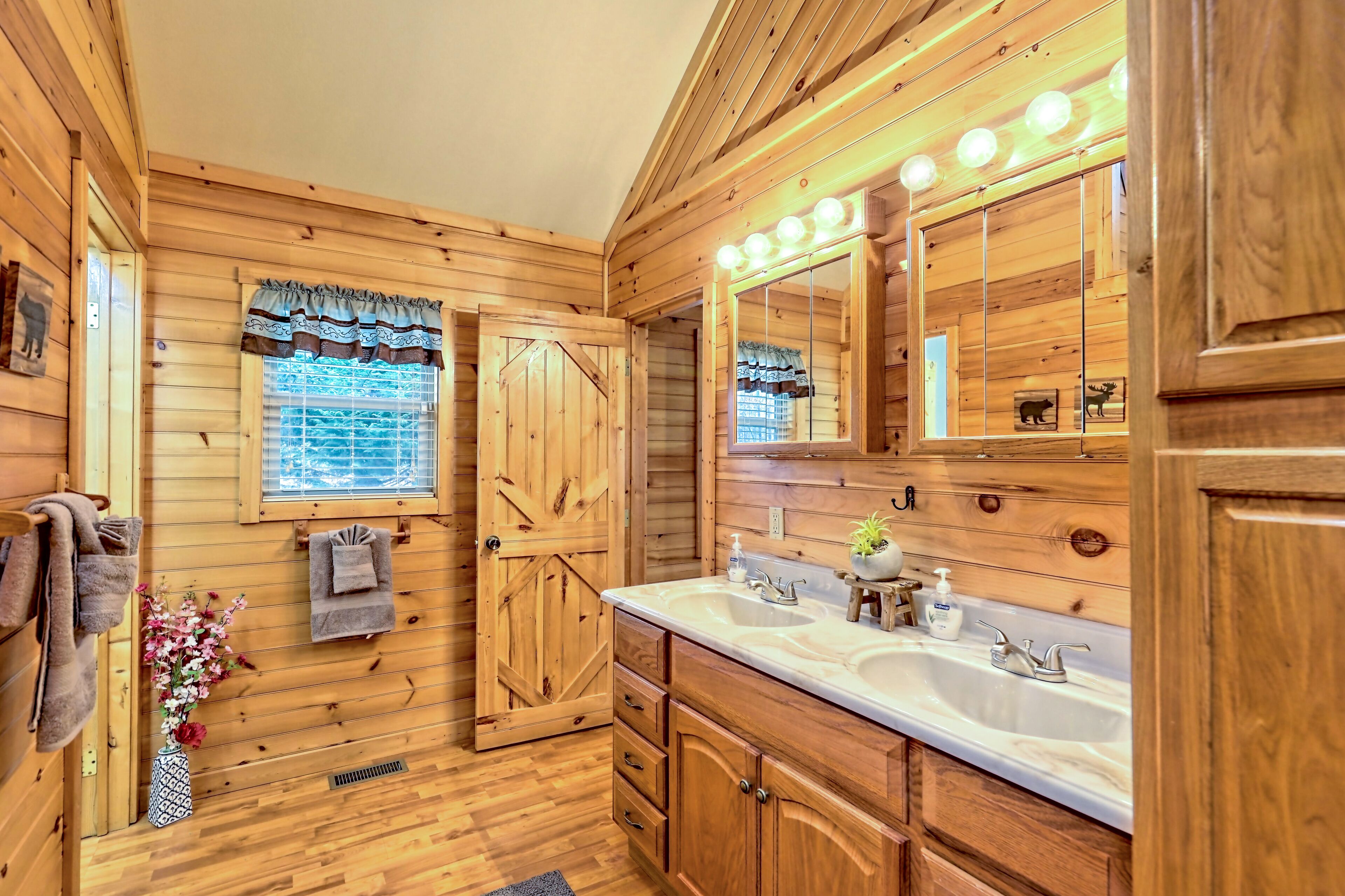 A wooden cabin bathroom with a large mirror, two sinks, wooden cabinets, and a window with a curtain.