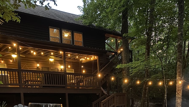 A cozy outdoor seating area with Adirondack chairs, a fire pit, and a wooden deck with string lights. - Cabin with hot tub firepit fireplace and game room area!near Helen  (Sautee Nacoochee)