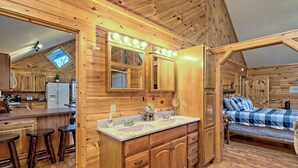 A cabin interior with a wooden interior, a kitchenette, a bathroom with a vanity and double sink, and a bed with a blue and white checkered bedspread. - Cabin with hot tub firepit fireplace and game room area!near Helen  (Sautee Nacoochee)