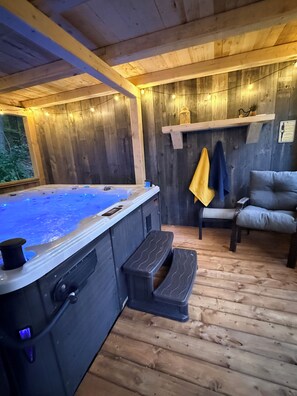 A wooden cabin interior with a hot tub, seating area, and a window overlooking greenery.