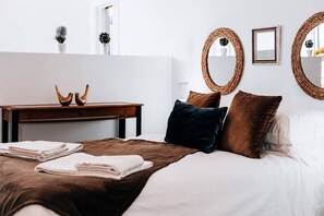 Una cama bien tendida con una manta y almohadas marrones, una mesita de noche de madera con dos objetos decorativos y dos espejos tejidos en la pared.