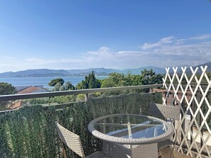A balcony with a wicker chair and a glass-top table, overlooking a body of water and distant hills.