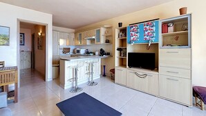 Een moderne keuken met een centraal eiland, barkrukken, een eethoek met een tafel, en een zithoek met een televisie en boekenkasten.