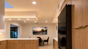 A modern kitchen with wooden cabinets, a central island, recessed ceiling lights, and a dining area with chairs.