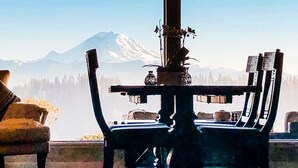 Uma área de refeições com mesa e cadeiras, um vaso com flores e vista para uma cordilheira através de grandes janelas.