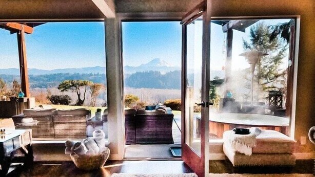 Interior moderno com uma grande janela que oferece vista para as montanhas. Há um espaço de estar aconchegante com um tapete e uma pequena mesa.