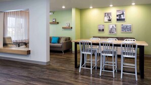Un espace repas moderne avec une table en bois et des chaises en métal, un canapé agrémenté d’un coussin bleu et une fenêtre ornée de rideaux légers.