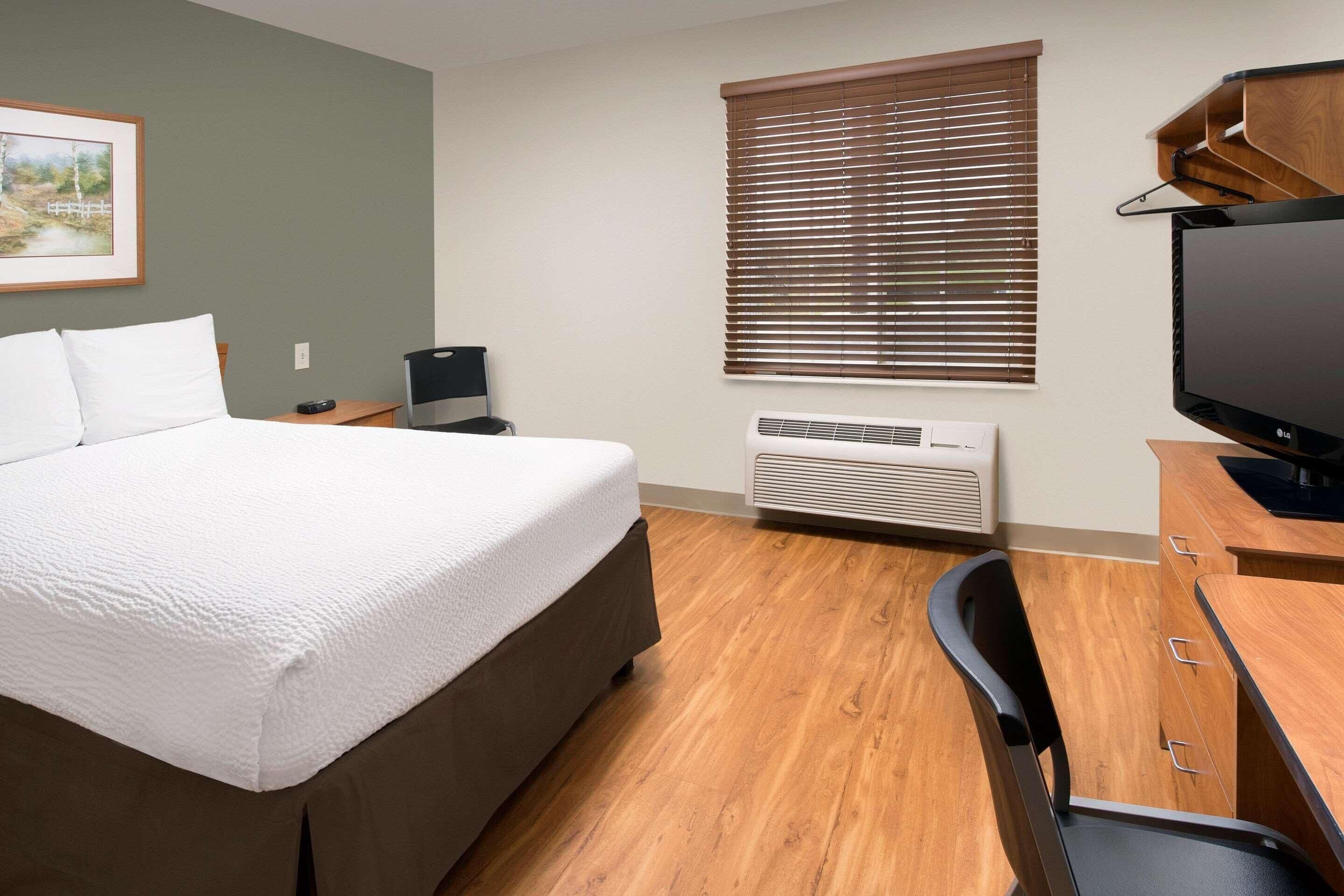 Quarto de hotel com cama, escrivaninha, cadeira, televisão e janela com persianas.