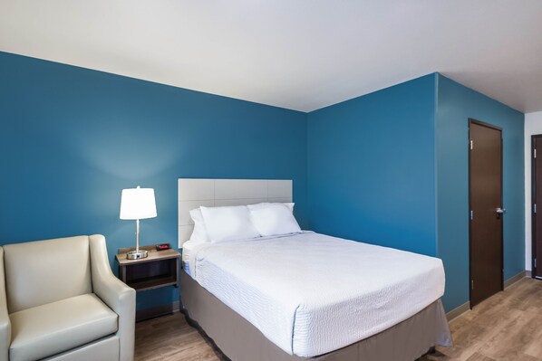 A hotel room with a bed, a nightstand, a lamp, a chair, and a door.