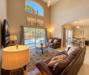 A spacious living room with a leather sofa, a coffee table, and a TV. There is a large window with a view of a swimming pool.