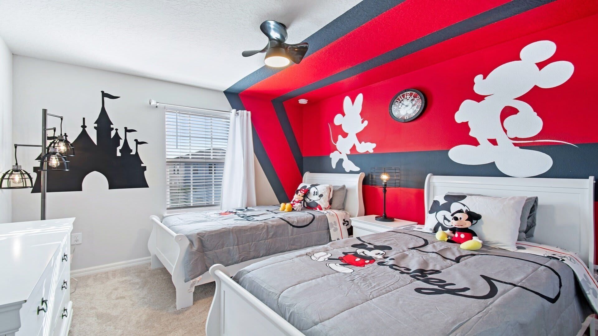 Un dormitorio con dos camas, cada una con ropa de cama temática de Mickey Mouse, un reloj en la pared y un adorno con silueta de castillo.