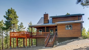 Une cabane moderne en bois, dotée d’une terrasse, d’une cheminée et d’un escalier menant à l’entrée principale.