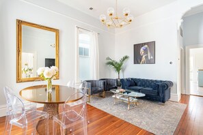 Una sala de estar con una mesa de comedor redonda, sillas transparentes, un sofá azul oscuro, una mesa de centro de vidrio y un espejo grande.