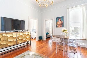 Un salon moderne doté d’un téléviseur à écran plat, d’un meuble en bois orné de motifs circulaires, d’un coin repas avec une table et des chaises en verre, et d’une grande fenêtre avec des rideaux.