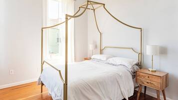 Una habitación con una cama con dosel, una mesita de noche con lámpara y un ventanal con cortinas.