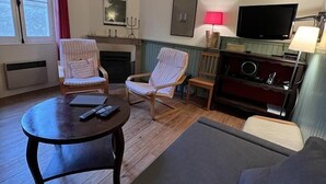 Una sala de estar con un sofá, dos sillones a rayas, una mesa de centro con mandos a distancia, un televisor, una chimenea y una ventana con cortinas.