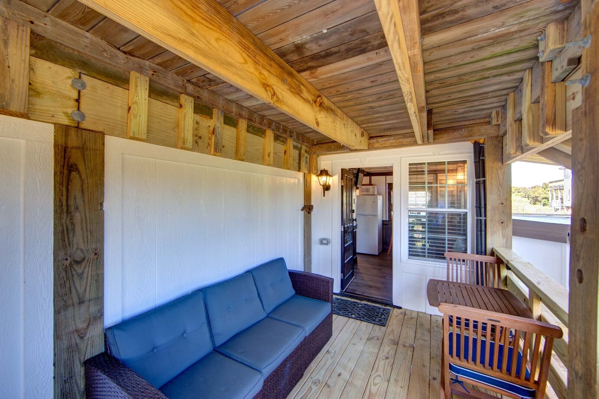 A rustic cabin interior with a wooden ceiling, a blue sofa, a wooden dining table, and a kitchen area visible through a doorway.