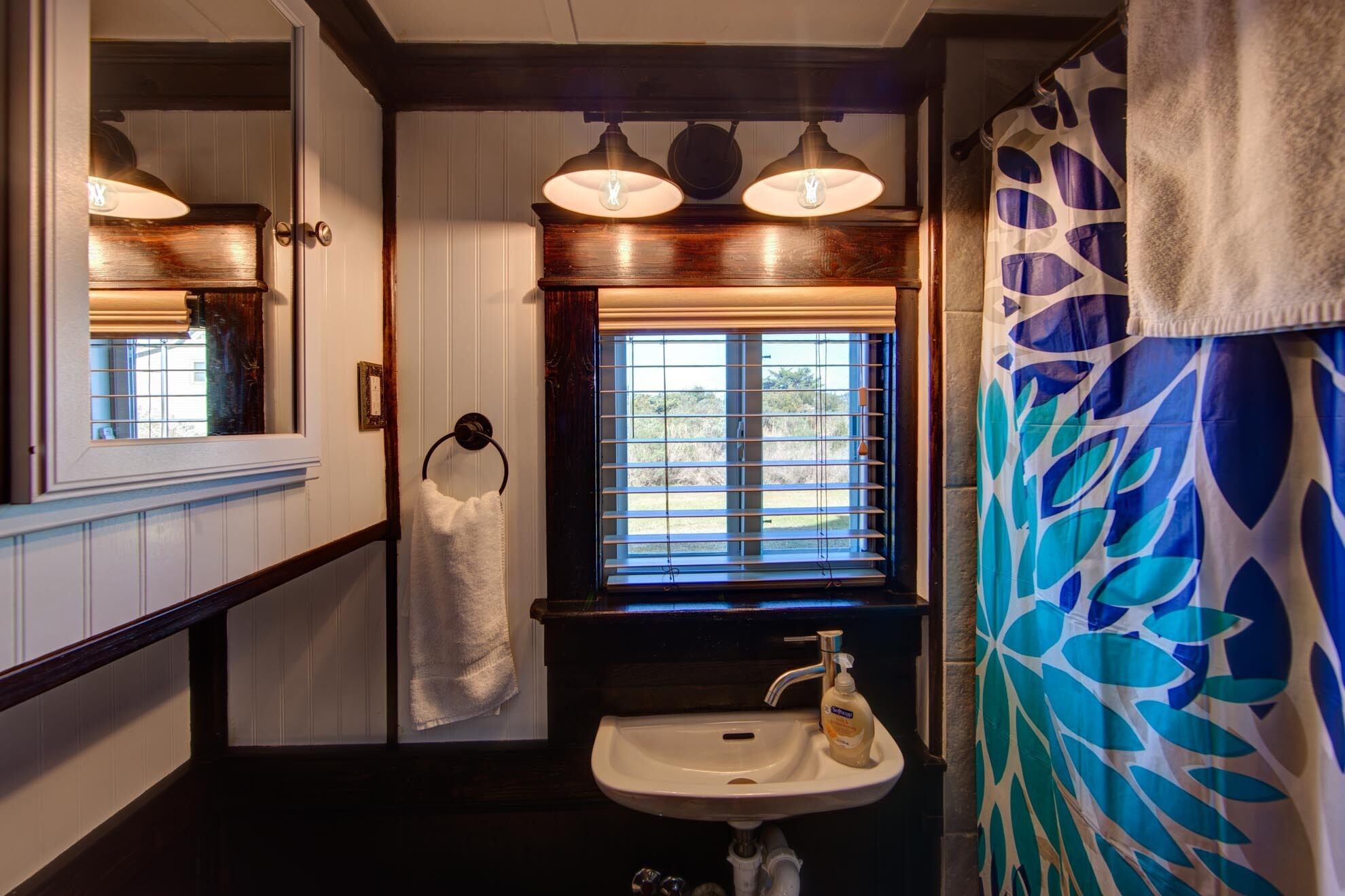 A bathroom with a white sink, a window with blinds, a towel hanging on a rack, and a shower curtain with a leaf pattern.