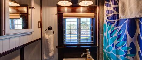 A bathroom with a white sink, a window with blinds, a towel hanging on a rack, and a shower curtain with a leaf pattern.
