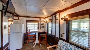 A compact kitchen with a refrigerator, microwave, and wooden cabinets. There is a dining area with a round table and two chairs. A patterned sofa is placed near a large window with blinds. - Historic suite with a private hot tub! (Avon)