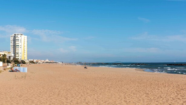 Une vaste plage de sable, avec un grand immeuble sur la gauche et un ciel bleu dégagé.