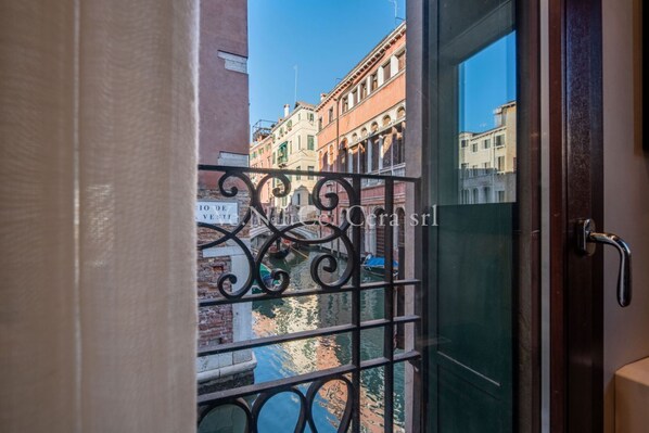 A view through a glass door with a metal railing, overlooking a canal with buildings in the background.