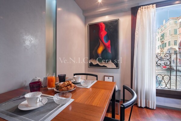 A dining area with a wooden table set for breakfast, a painting on the wall, and a view of buildings through a window.