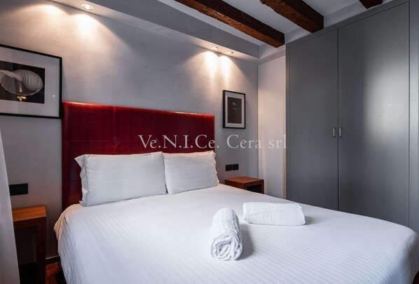 A hotel room with a large bed, a red headboard, white bedding, and a neatly folded towel on the bed.