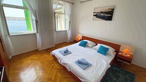 A hotel room with a wooden floor, a bed with white linens and blue pillows, two bedside tables with lamps, a window with sheer curtains, and an air conditioning unit on the wall. - Holiday apartment with two bedrooms and sea views (Jadranovo)