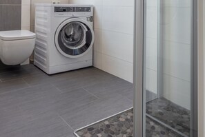 A modern bathroom with a white toilet, a front-loading washing machine, and a shower area. - 50062 "Leev mare" - Sophisticated hideaway in Altfunnixsiel: stylishly modern (Wittmund)