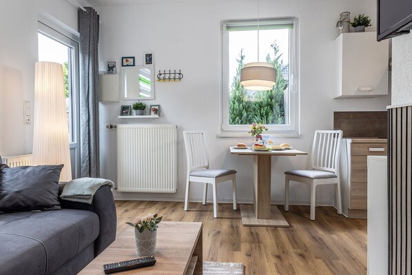 Un espace de vie compact comprenant un canapé, une table à manger avec des chaises, un radiateur, une fenêtre donnant sur un espace verdoyant et un téléviseur.