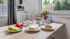 Une table dressée pour le petit-déjeuner, avec du fromage, des tomates, des œufs et des viennoiseries, placée près d’une fenêtre donnant sur une clôture et de la verdure.