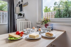 Aamiaispöytä, jossa on juustoa, tomaatteja, kananmunia ja leivonnaisia, ja pöytä on sijoitettu ikkunan lähelle, josta avautuu näkymä aitaan ja vihreään maisemaan.