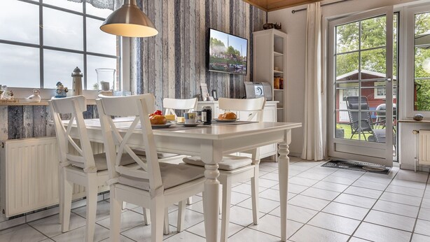 A dining area with a white table and chairs, a pendant light, and a television on the wall. - Vacation home 50059 - Arrive in Altfunnixsiel and feel at home: Quiet (Wittmund)