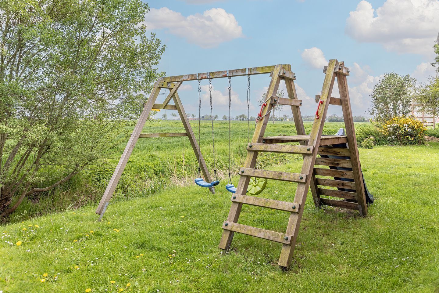Une balançoire en bois installée dans un champ verdoyant, avec des arbres et un ciel dégagé.