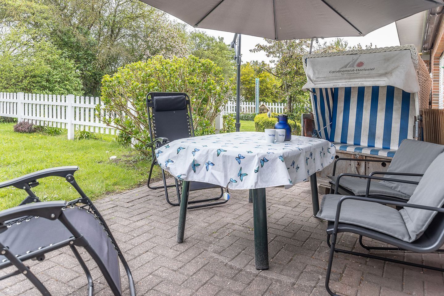 An outdoor patio with a table covered by a white tablecloth with blue butterfly patterns, surrounded by metal chairs.