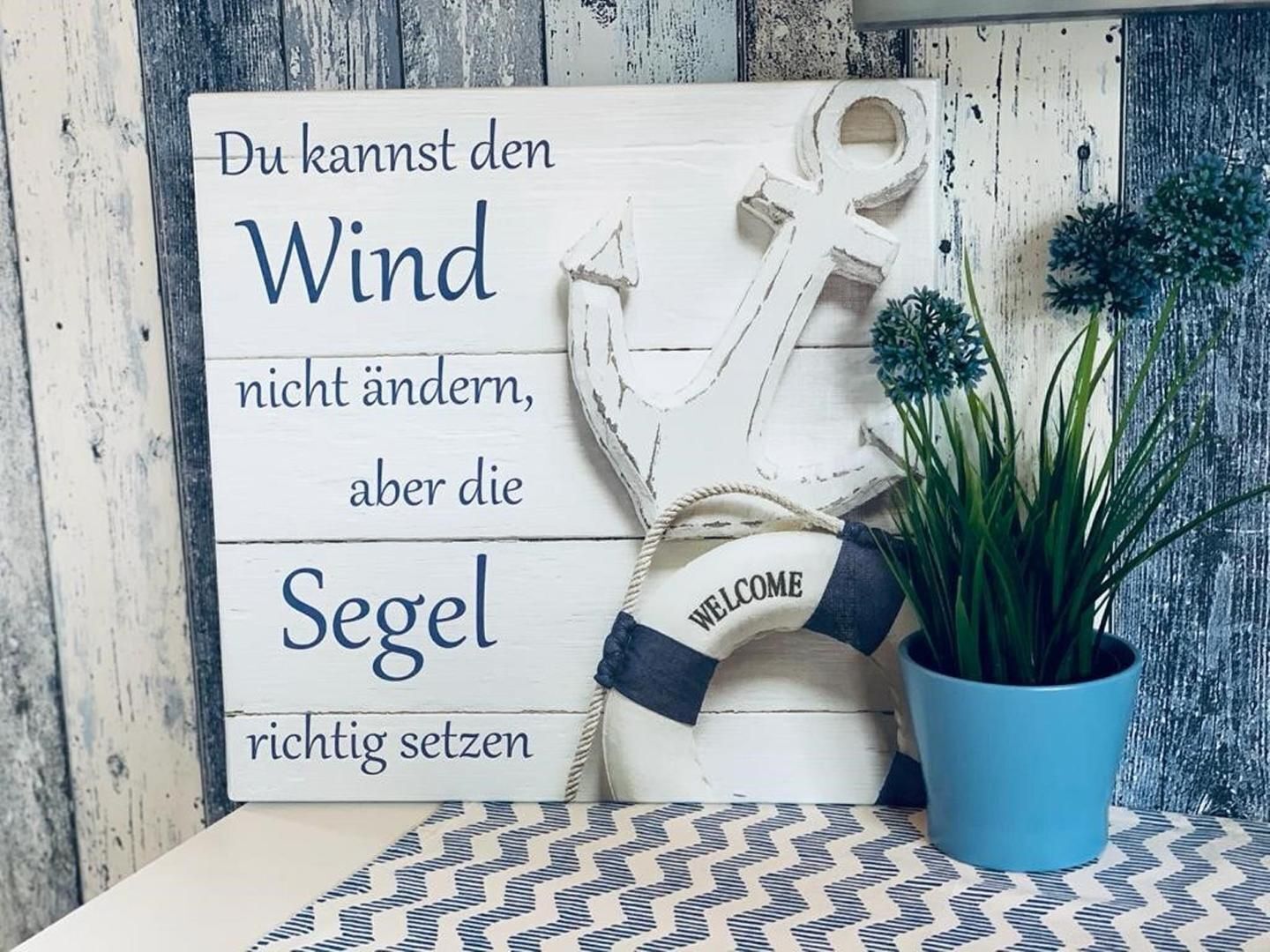Ein Holzschild mit Schriftzug, ein Rettungsring und ein Blumentopf auf einer gemusterten Fläche.
