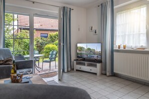A living room with a TV, a sofa, a dining table, and chairs. There is a view of a garden and a building outside the window. - Vacation home 50007 - pure comfort and relaxation: the semi-detached house with (Wittmund)