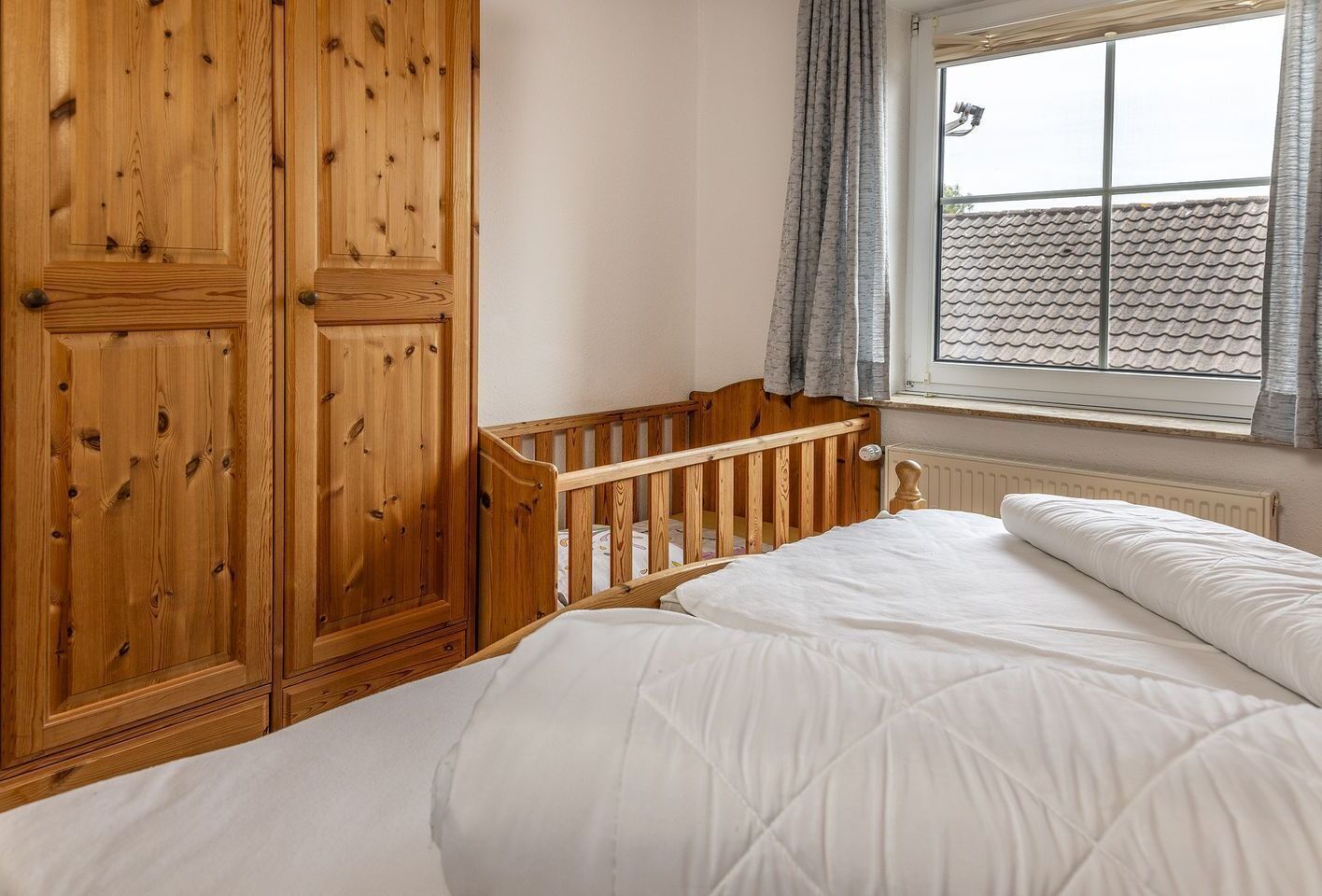 Un dormitorio con un armario de madera, una cuna, una cama con ropa blanca y una ventana con cortinas.