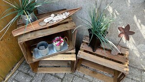 Un décor extérieur rustique, avec des caisses en bois disposées dans un coin, contenant divers objets tels que des plantes, une étoile décorative en métal et un plateau avec des fleurs.