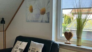 Une pièce avec un canapé en cuir noir, deux coussins ornés de l’inscription « Coast 53 », une œuvre d’art encadrée représentant des fleurs, une grande plante verte et une décoration en forme de cœur posée sur un rebord de fenêtre.