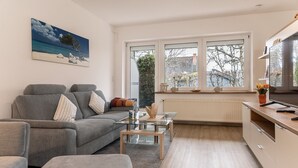 A modern living room with a grey sofa, a glass coffee table, and a view of the outside through large windows. - Open and flooded with light: the first floor apartment with its 62 square meters was brightly (Wittmund)
