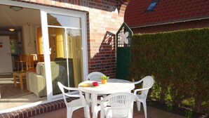 Eine Terrasse mit einem runden Tisch, weißen Plastikstühlen, einem Blumentopf und einem kleinen Tisch mit Schüssel und roter Tasse.