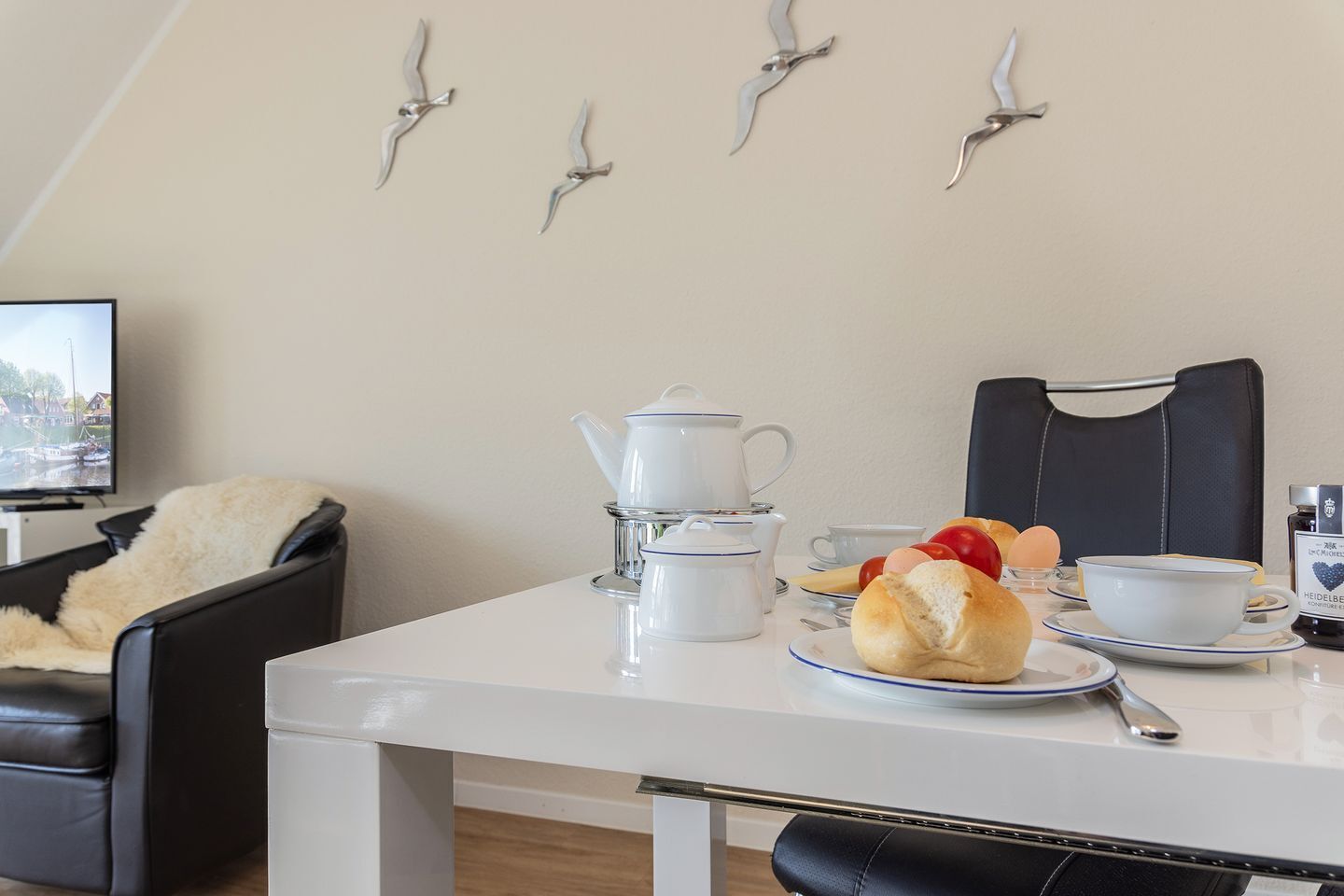 Een eetgedeelte met een witte tafel gedekt voor het ontbijt, inclusief brood, eieren en een theepot. Er staat een zwarte lederen stoel en er is een televisie op de achtergrond te zien.