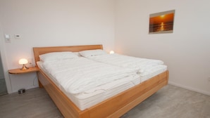 A neatly made bed with white linens in a room with a wooden headboard. There is a nightstand with a lamp and a wall-mounted thermostat. - Bright and open: The homely apartment, which was completed in 2017 (Wittmund)