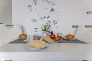 Une table blanche est dressée pour le petit-déjeuner avec du pain, du fromage, des tomates et une tasse de café. Sur le mur, on peut admirer une décoration représentant des illustrations de café et de thé.
