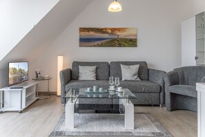 Un salon moderne comprenant un ensemble de canapés, une table basse en verre, un téléviseur à écran plat et un tableau représentant un paysage côtier.