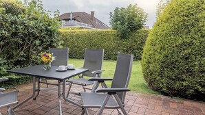 Eine Terrasse im Freien mit Tisch und Stühlen, einer Vase mit Blumen und einem gepflegten Heckenabschnitt.