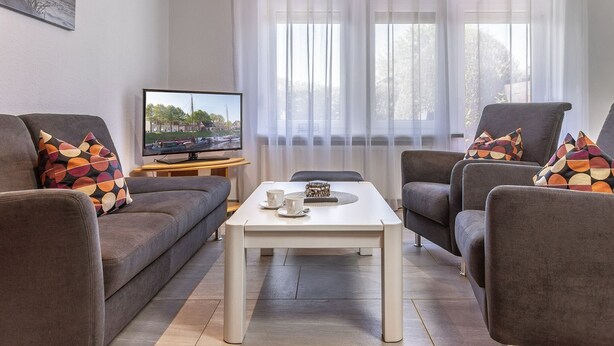 Ein Wohnzimmer mit einer Couch, Sesseln, einem Couchtisch, einem Fernseher und einem Fenster mit durchsichtigen Vorhängen.