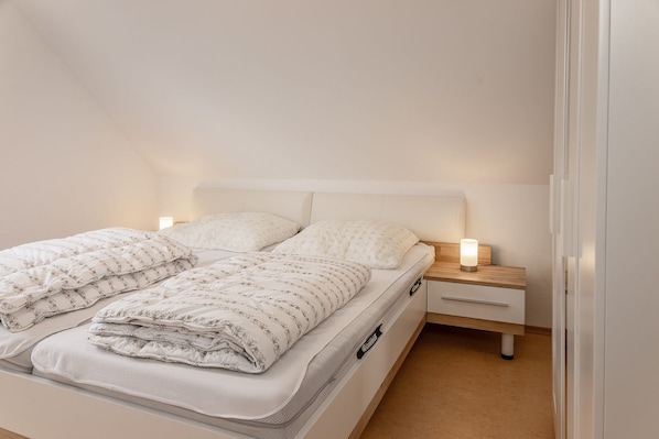 Ein ordentlich bezogenes Bett mit einem weißen Kopfteil, ein Nachttisch mit Lampe und ein Holzbeistelltisch.